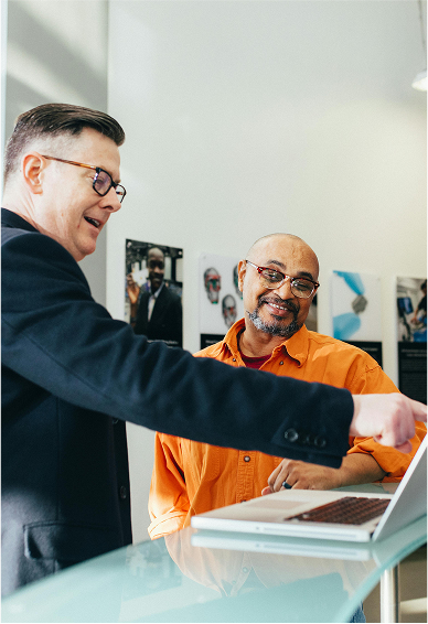 Two men look at a laptop screen on a glass countertop while one of them points at something on the screen.